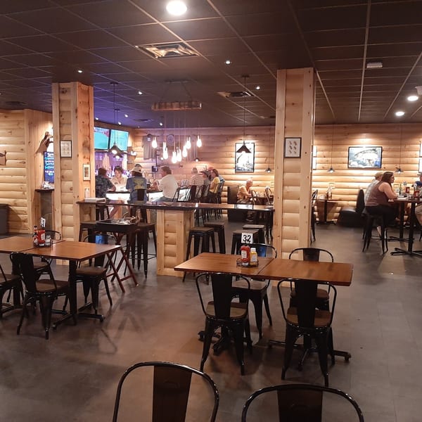A rustic restaurant interior with wooden walls, dark tables, and patrons dining and socializing.