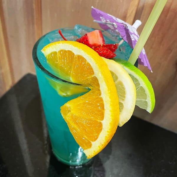 Blue cocktail garnished with orange, lemon, lime slices, strawberries, a cherry, and a paper umbrella.